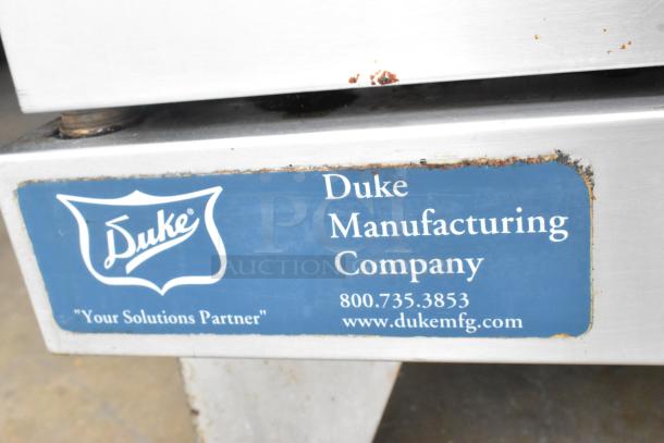 Duke Manufacturing label on a stainless steel convection oven, showing logo, contact info, minor rust on corners.