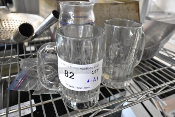 Lot of glass mugs and metal items, including two clear mugs with "82 QTY" labels, displayed on metal shelving.