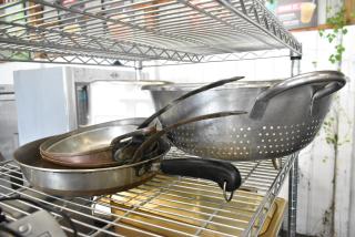 Seven metal kitchen items including a large colander and various skillets with handles, stacked on a wire shelf.