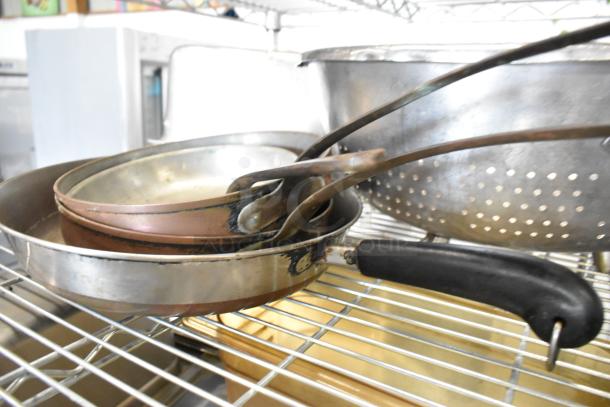 7 assorted metal items, including skillets with wooden handles and colander, on a wire rack, showing moderate wear.