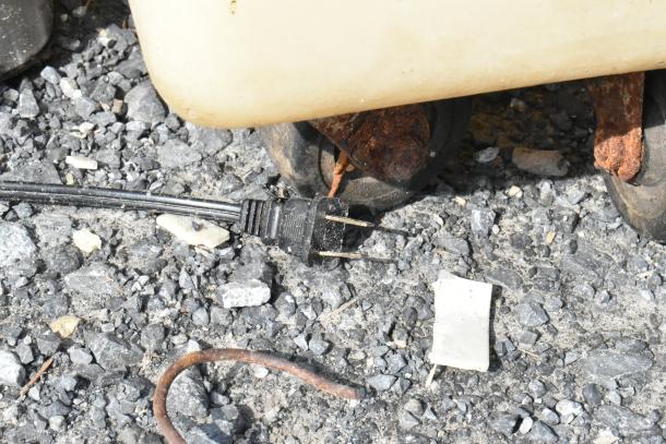 A close-up view of an electrical plug on gravel, partially obscured by rusted equipment, with no coffee machine visible.
