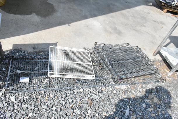 Lot of 6 various wire shelves, mixed sizes and colors, stacked outdoors on gravel. Some rust and wear visible.