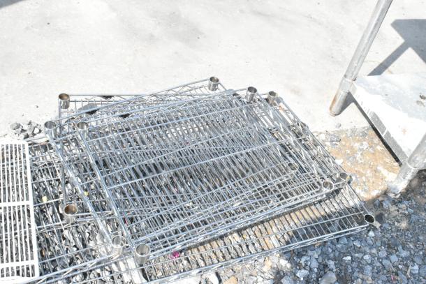 Lot of 6 various metal wire shelves in used condition, stacked on a gravel surface, showing corner posts and grid pattern.