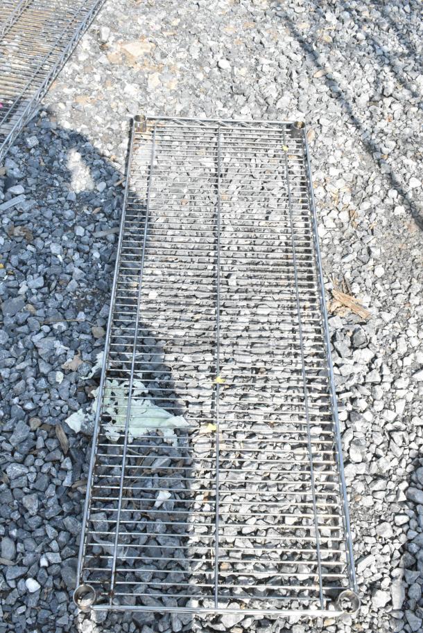Lot of 6 metal wire shelves on gravel, showing minor wear. Ideal for storage organization. No visible markings.