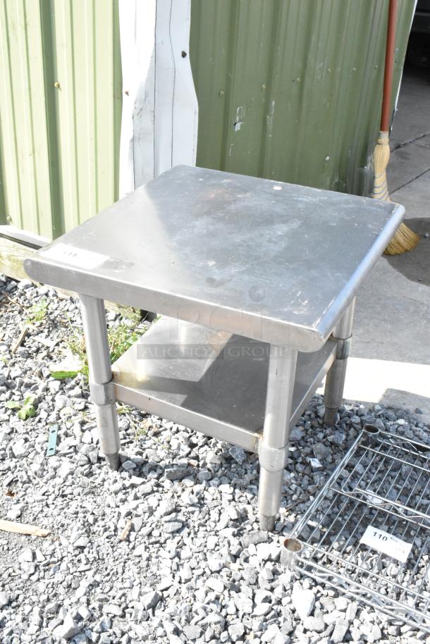 Stainless steel commercial equipment stand with metal under shelf, outside on gravel. Sturdy condition, ideal for heavy-duty use.