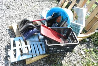 Pallet lot with snow shovels, poly bucket, metal shelf, and misc. items on gravel, outdoor setting.