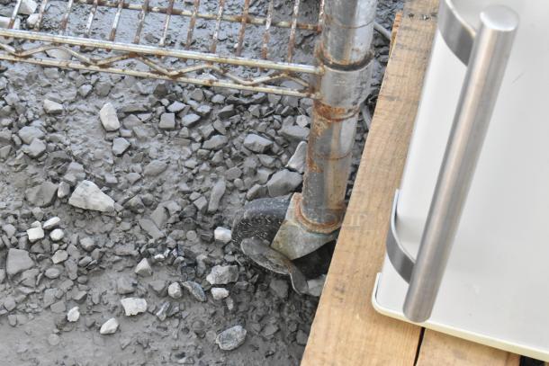 Stainless steel table with wire under shelf on commercial casters, showing minor rust on legs, set on gravel surface.
