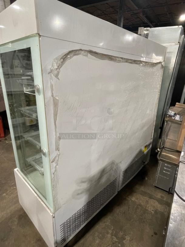 Commercial white glass refrigerated bakery merchandiser with multi-tier shelves and double glass sliding doors on casters. Slight wear visible.