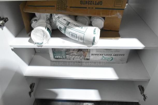 White and red counter with POS monitor, showing storage shelves. Contains packaged cups and a bakery display case.