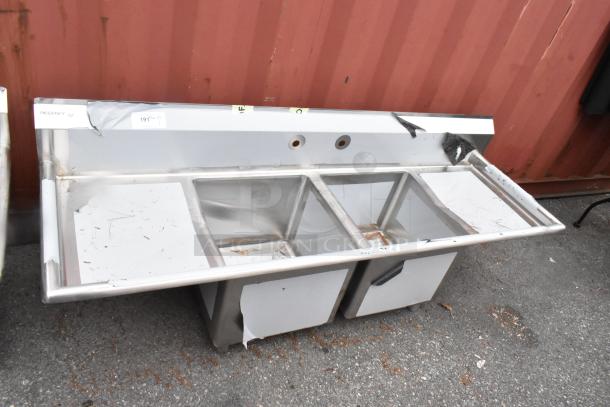 Brand new Regency 600S21818218 stainless steel 2-bay sink with dual drain boards. Visible scratches and dents. Tags present.