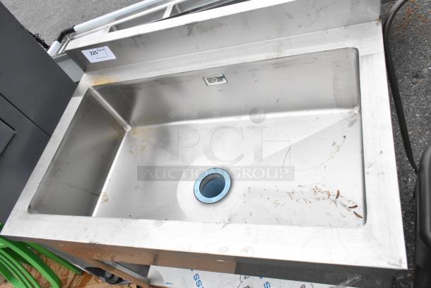 Stainless steel sink bay with under shelf, showing minor wear. Features drainage hole and back splash panel. Auction tag visible.