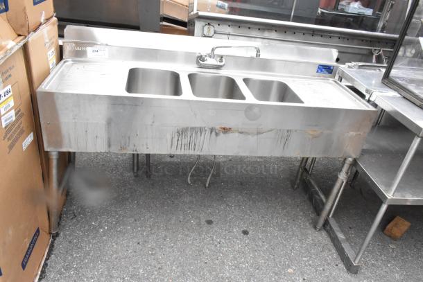 Stainless steel 3 bay bar sink with dual drain boards, faucets, and handles. Visible wear and marks on front.