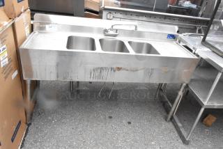 Stainless steel 3 bay bar sink with dual drain boards, faucets, and handles. Visible wear and marks on front.