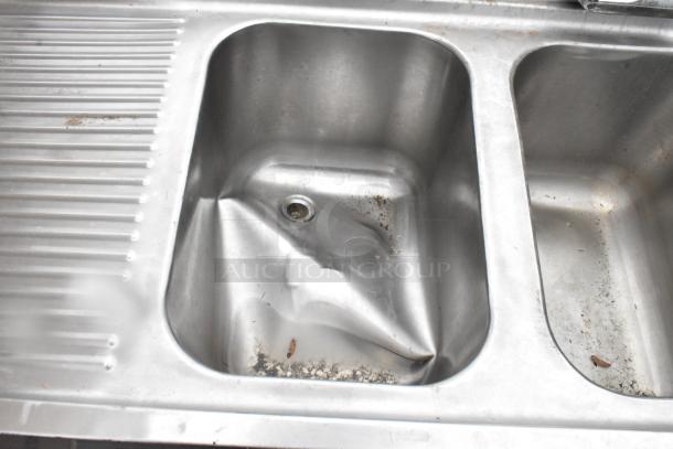 Stainless steel 3 bay bar sink with visible dents, dual drain boards, faucets, and handles. Some grime present.