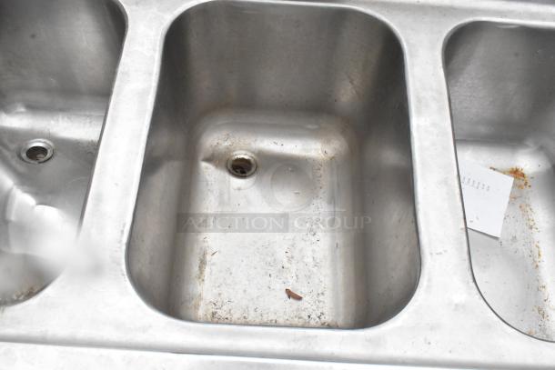 Stainless steel 3-bay bar sink with visible wear, dual drain boards, and faucets. Ideal for commercial use.
