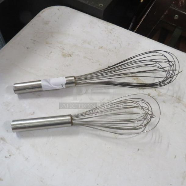 Two stainless steel wire whisks of assorted sizes on a table, featuring metal handles. Slightly used condition.