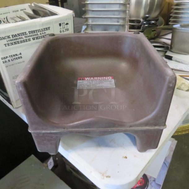 Brown booster seat, plastic material, slight wear. Warning label on seat surface. Measures approximately 15x15 inches.