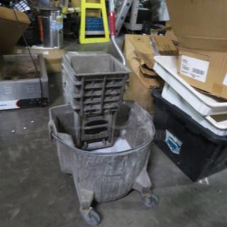 Gray mop bucket with attached wringer, four wheels for mobility, used condition, ideal for commercial cleaning needs.