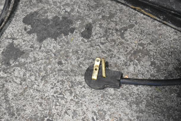 Close-up of a damaged electrical plug on concrete. The prongs are bent and crooked.