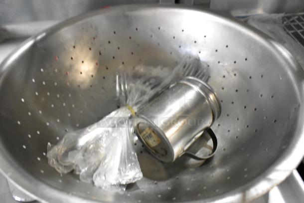 Lot of kitchen items including a metal colander, plastic-wrapped utensils, and a metal baking pan.