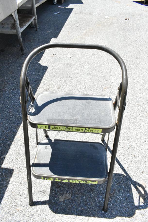 Metal folding step ladder with two steps, safety labels instruct folding when not in use, in used condition.