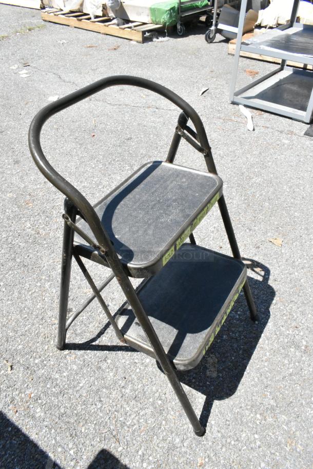 Folding step ladder with two black steps and sturdy metal frame. Surface shows light wear, suitable for home use.