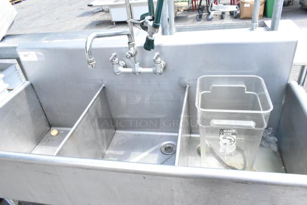 Stainless steel 3 bay sink with dual-handle faucet, spray nozzle attachment, and visible wear. Includes a plastic bin with label.