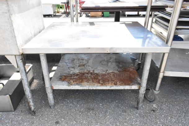 Stainless steel equipment stand with metal under shelf, showing signs of rust and wear. Features casters for mobility.