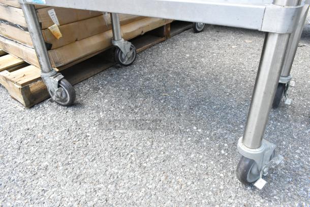 Stainless steel table with metal under shelf on commercial casters, shown on concrete surface. Rugged, industrial design.