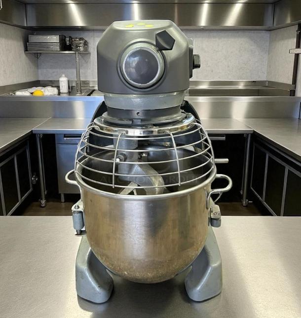 Commercial stand mixer with metal bowl, protective cage, and control knobs. Appears in good condition. Suitable for heavy-duty use.