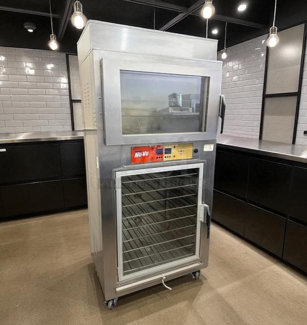 Nu-Vu commercial oven with glass front, metal exterior, multiple racks, on caster wheels, control panel with brand label.