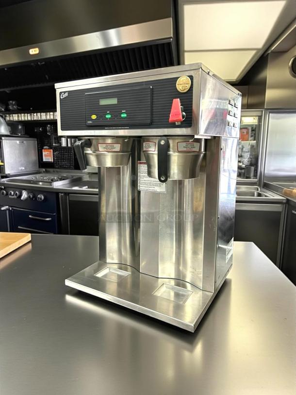 Commercial coffee brewer with dual stainless steel urns, digital control panel, and visible brand markings. Ideal for high-volume use.