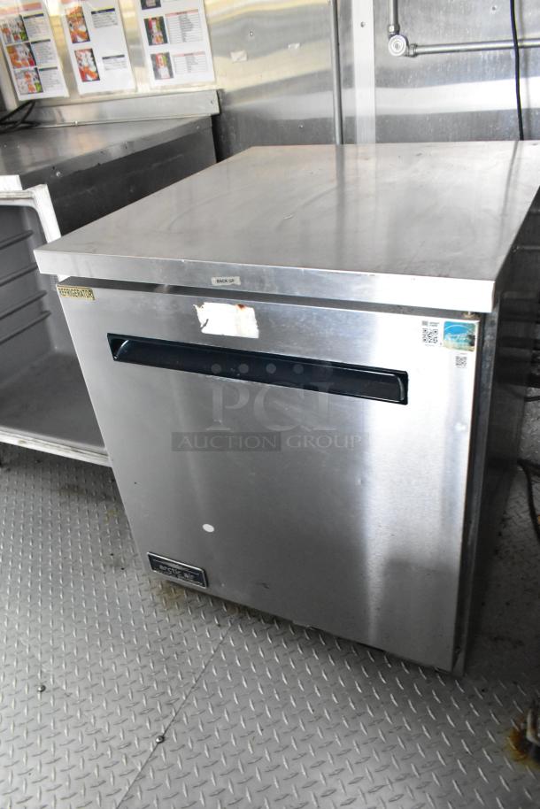 Stainless steel refrigerator in former Popeyes food trailer, featuring a label marked "Refrigerator" and handle on front.