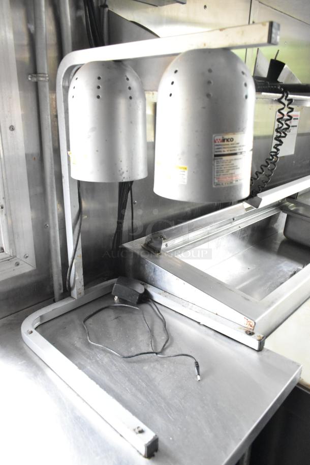 Stainless steel prep area in a former Popeyes food trailer, featuring a heat lamp and cable, with visible wear.