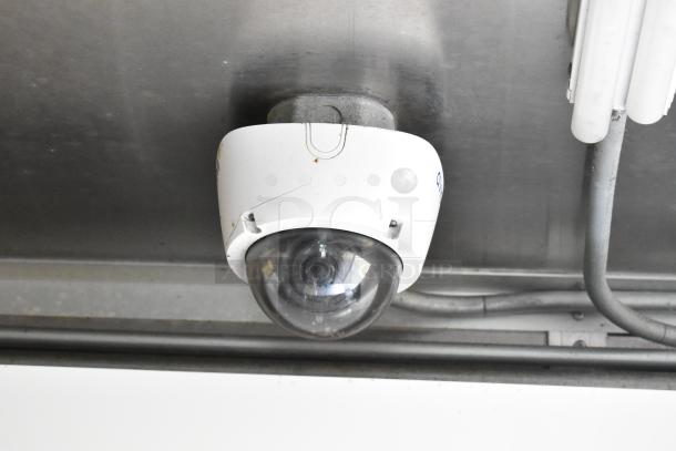 A white security camera mounted on a metal ceiling inside a trailer kitchen.