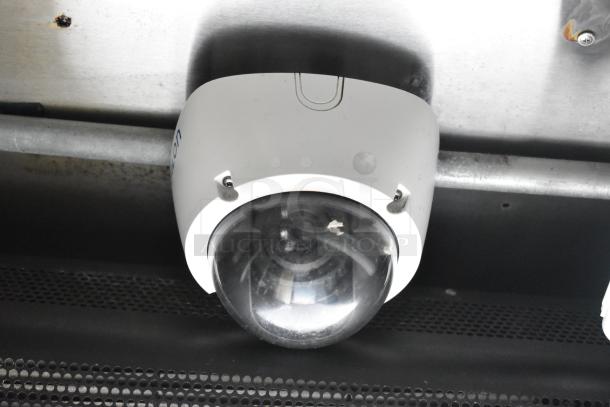 Close-up of a white dome-shaped security camera mounted on a ceiling with a metallic pipe nearby.