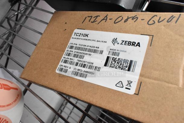 Cardboard box with a Zebra TC210K label showing details like part number, serial number, and manufacture date.