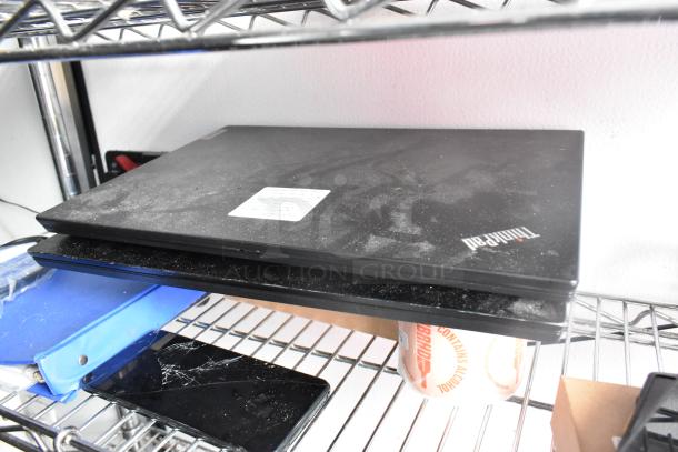 Laptop on metal shelving in a warehouse. The laptop appears dusty, with a visible Lenovo logo. Nearby, a blue binder and a cracked smartphone screen are also on the shelf.