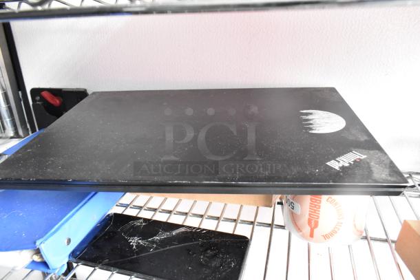A black cutting board with a white logo on a metal shelf near a blue folder and a partially visible canned beverage.