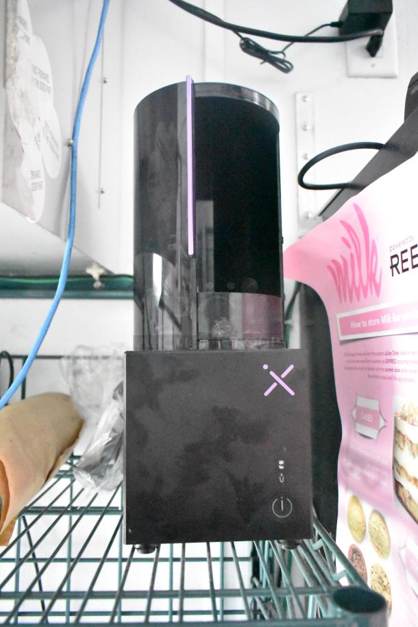 Sleek black appliance with vertical LED, positioned on a metal shelf. Nearby, a pink Milk Bar product guide.