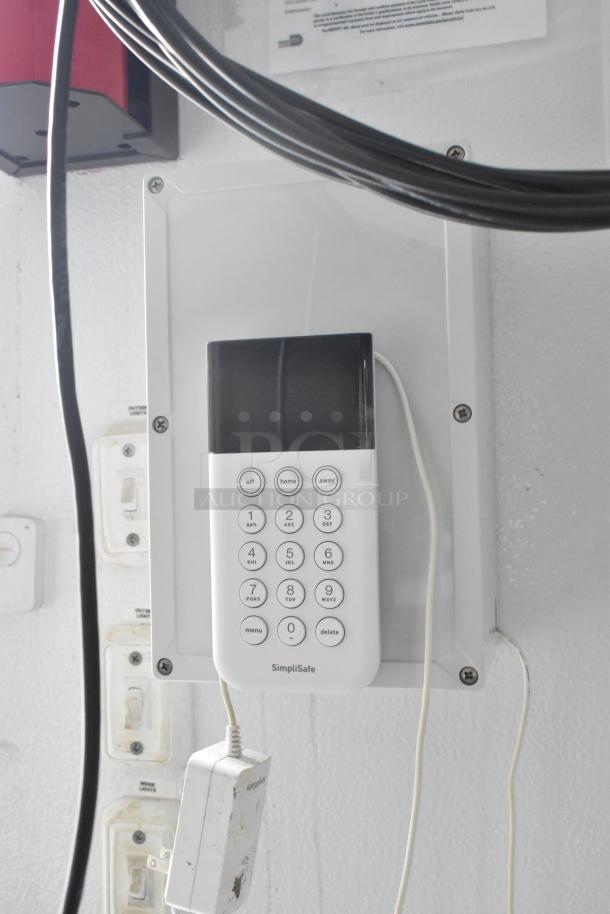 SimpliSafe keypad on wall with wiring. White plug labeled "SimpliSafe." Various wall switches visible nearby.