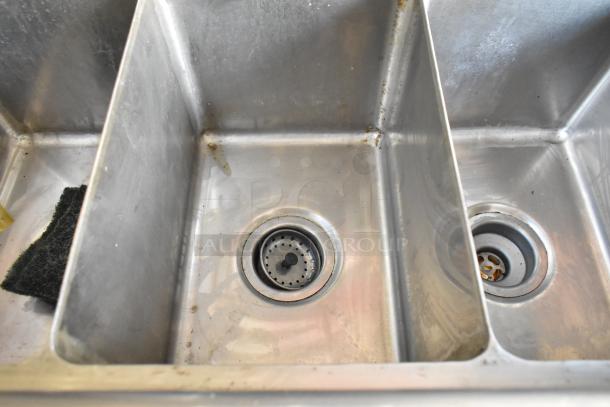 Three-compartment stainless steel sink inside a food trailer, slightly used with visible cleaning sponge and minor wear marks.
