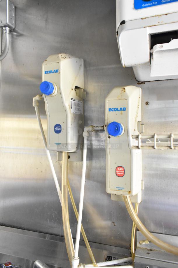 Close-up of Ecolab dispensers in a food trailer kitchen, marked for liquid detergent and multi-quat sanitizer, with hoses attached.