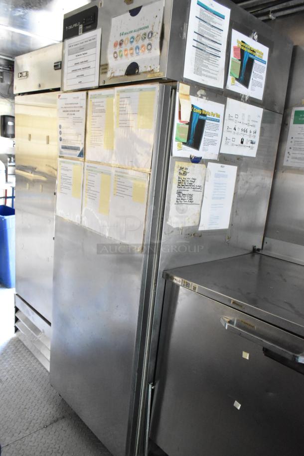 Mobile kitchen interior showing stainless steel coolers and prep tables, with various informational labels and notices attached.