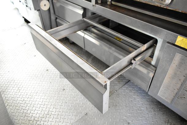 Mobile kitchen interior with open stainless steel drawer, featuring a grease hood and appliances. Textured metal flooring visible.