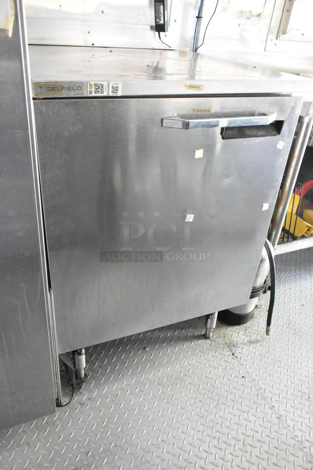 Stainless steel commercial fridge in food trailer, labeled Delfield, good condition, part of fully outfitted mobile kitchen.