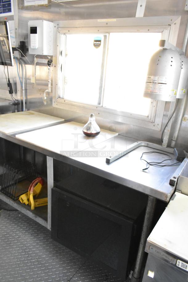 Fully outfitted 2019 food trailer kitchen interior, featuring stainless steel prep tables, window, and countertop lamp.