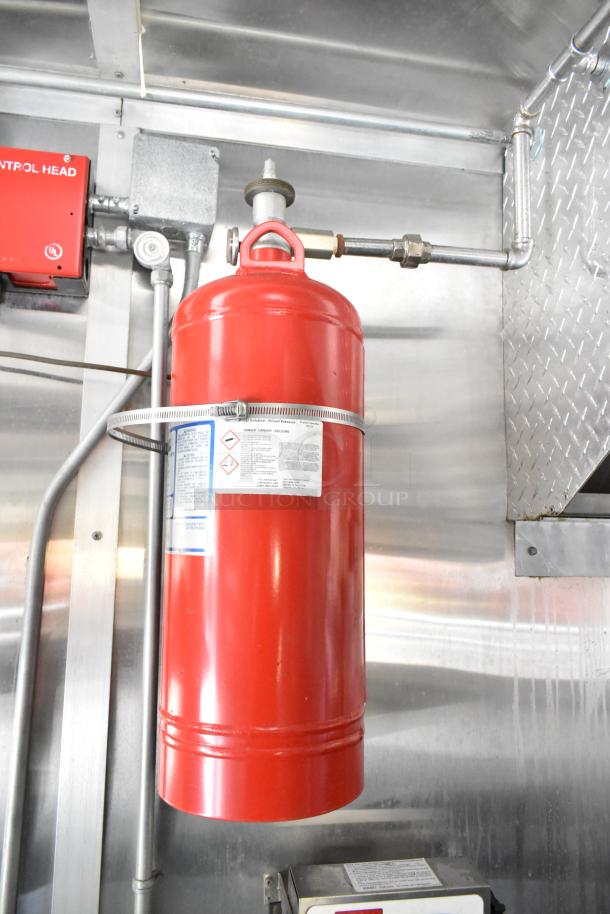 Red fire suppression system cylinder in a food trailer, mounted on a stainless steel wall with visible control head and piping.