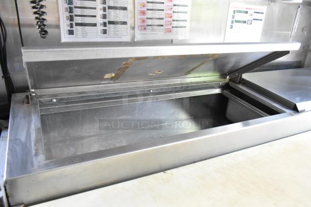 Stainless steel food trailer prep station with open lid, showing ample storage space. Includes menu chart on wall.