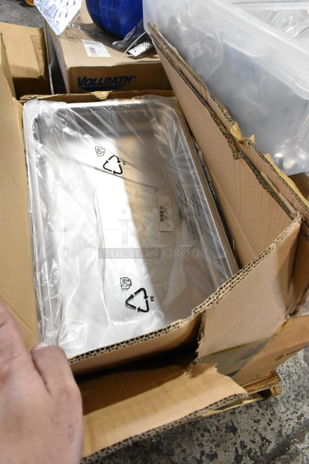 Cardboard box containing brand new metal trays wrapped in plastic, labeled with recycling symbols. Vollrath box visible.